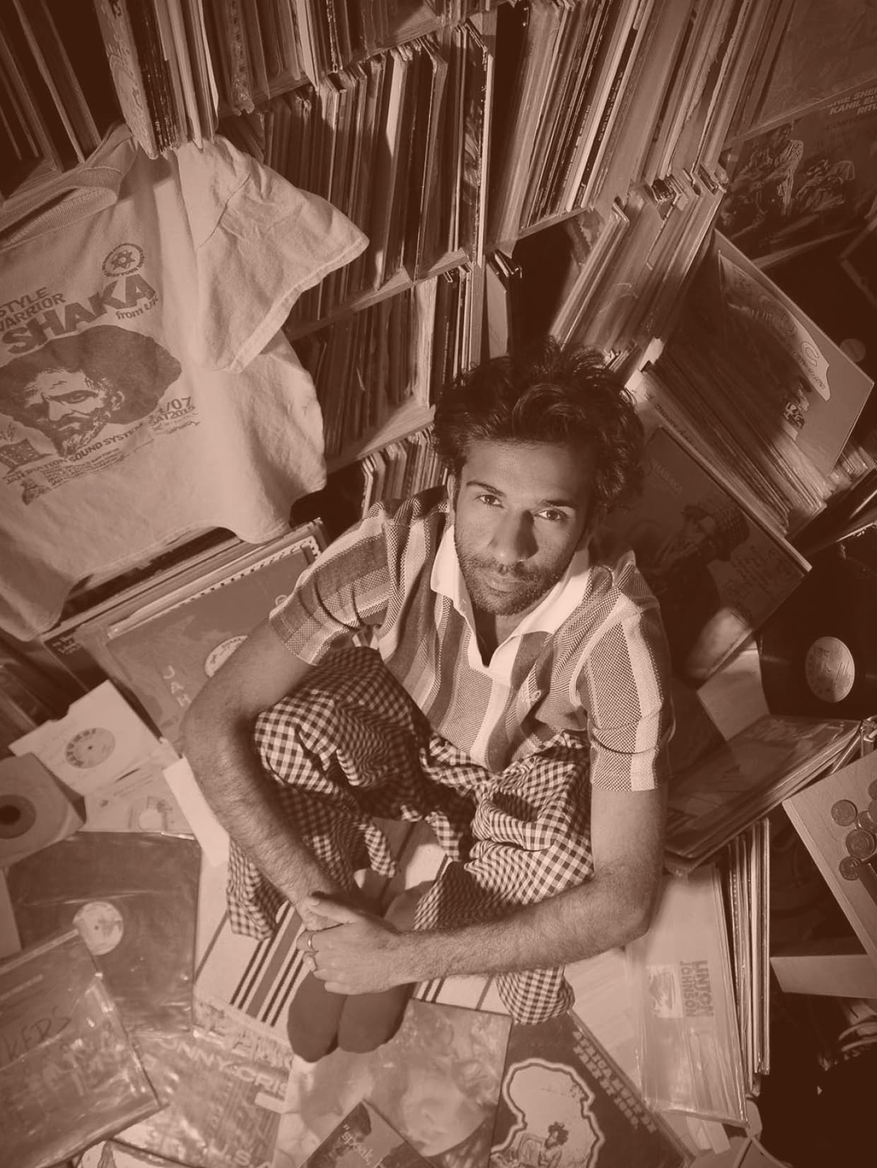 A portrait of a Haseeb Iqbal sitting among a large vinyl record collection, surrounded by shelves and stacks of records in a home music archive, looking up at the camera.